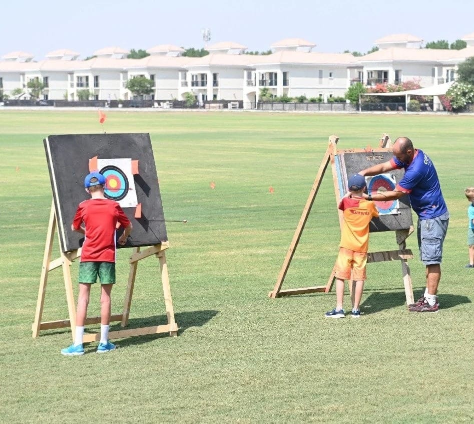 An outdoor archery session on a lawn, two children seen from behind at their targets with a coach.