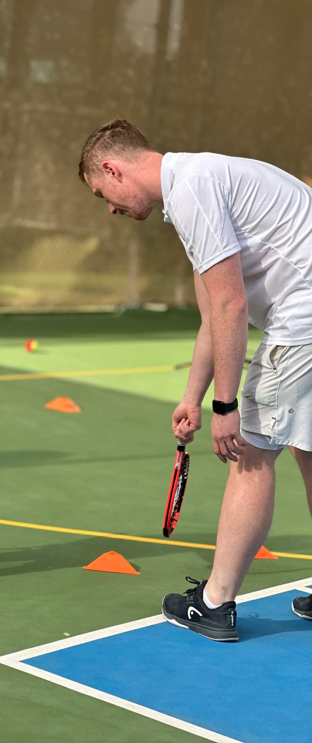 An adult coach bending with a racket over a line of orange cones on a green court.