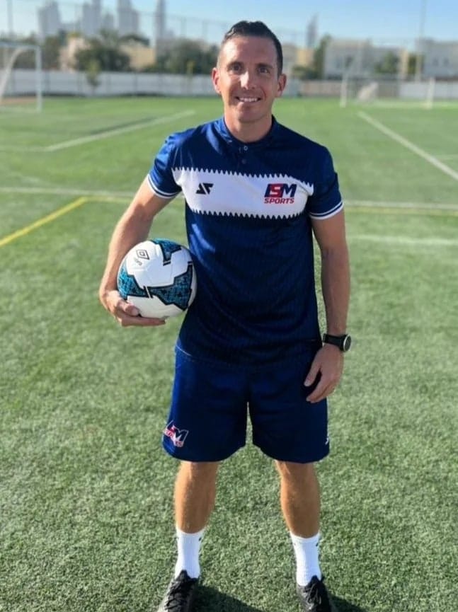 An adult coach in ISM Sports kit holding a football on artificial turf.