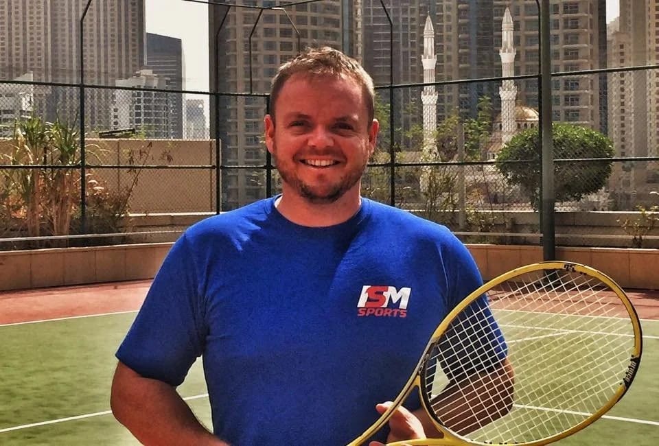 An adult in an ISM Sports shirt holding a racket, towers behind.