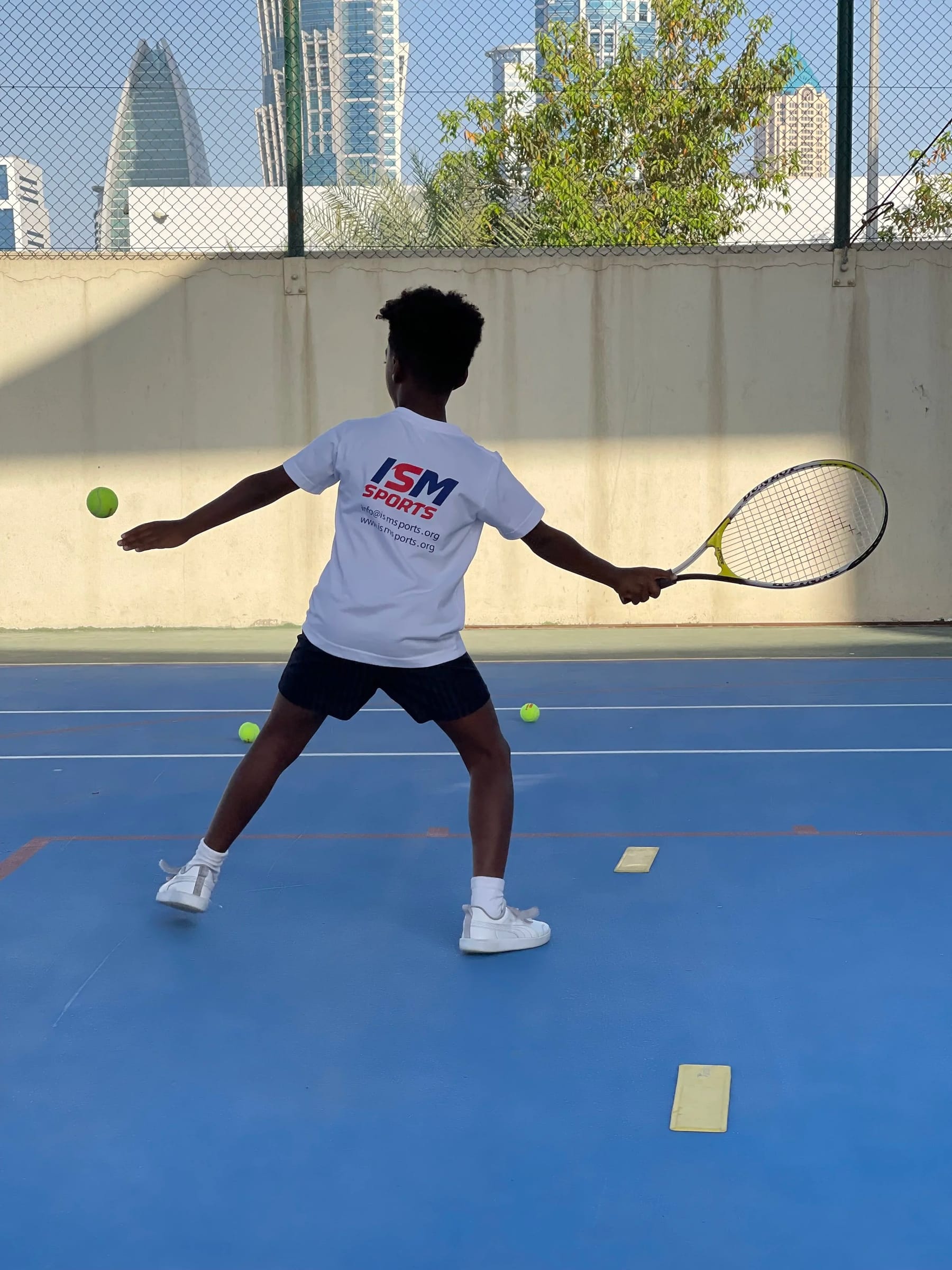 A child in an ISM Sports T-shirt, seen from behind, playing tennis on a blue court with the Dubai skyline beyond.