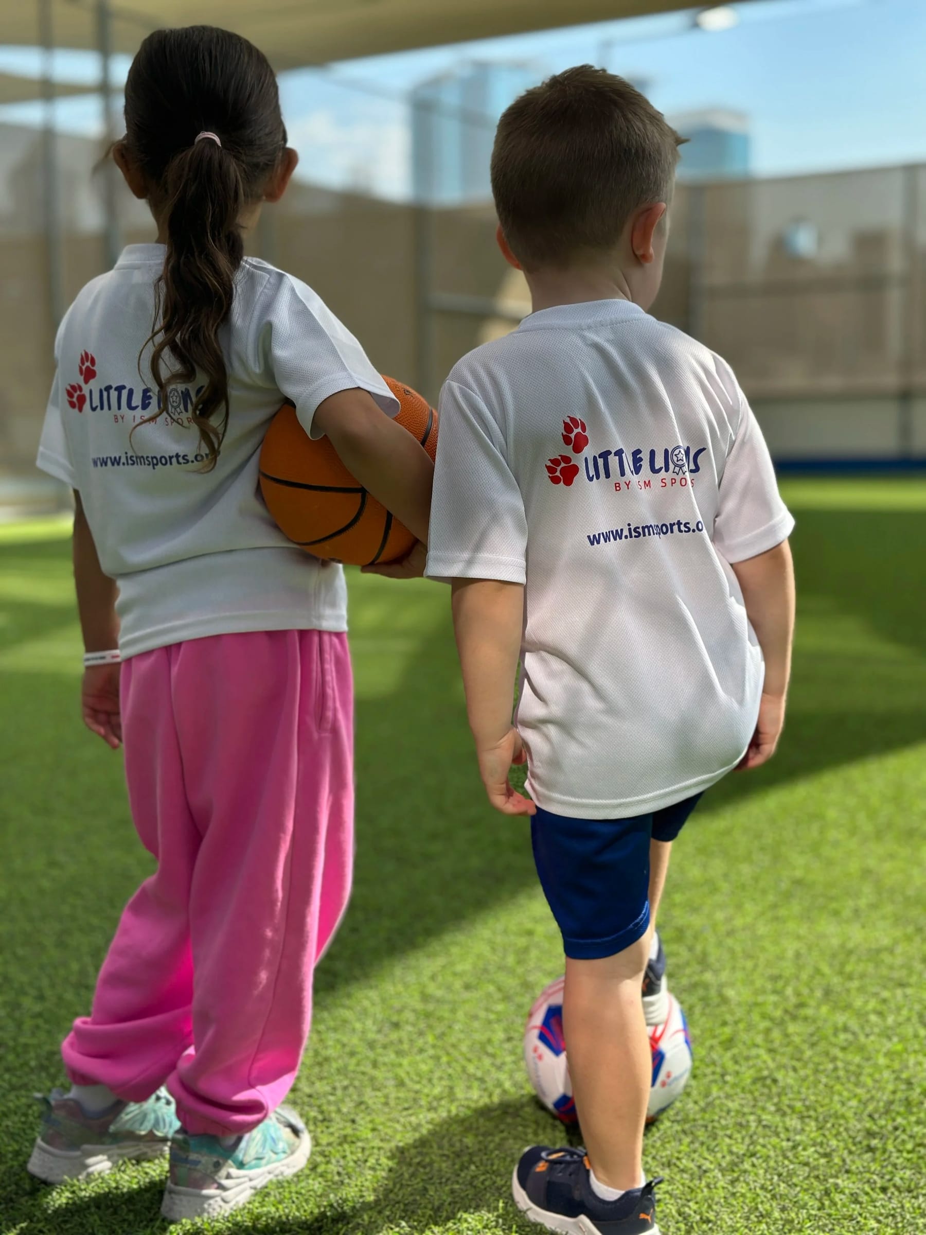 Two small children seen entirely from behind in Little Lions shirts, with a basketball and a football.